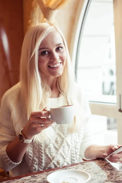 Una joven y hermosa mujer sostiene una taza de capuchino caliente. Una joven está sentada en un acogedor café bebiendo capuchino y hablando por teléfono. La chica está bebiendo cacao. El invierno. Acogedor otoño . — Foto de Stock