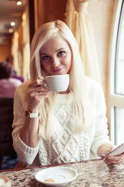 Una joven y hermosa mujer sostiene una taza de capuchino caliente. Una joven está sentada en un acogedor café bebiendo capuchino y hablando por teléfono. La chica está bebiendo cacao. El invierno. Acogedor otoño . — Foto de Stock