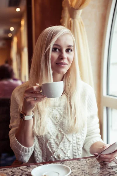 Una giovane bella donna sta tenendo una tazza di cappuccino caldo. Una giovane donna è seduta in un accogliente caffè a bere cappuccino e parlare al telefono. La ragazza sta bevendo cioccolata. Inverno. Autunno accogliente . — Foto Stock
