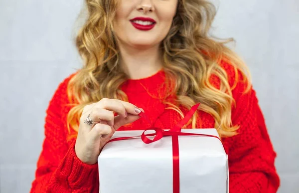 Ritratto di una giovane bella donna. Una giovane donna con un maglione rosso e un rossetto rosso regge un regalo. Trucco. Vacanze. San Valentino. Natale. Anno nuovo . — Foto Stock