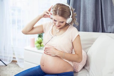 Genç güzel hamile kadın evde dinleniyor. Genç hamile kadın müzik dinliyor. Güzel bir hamile kadın portresi. Bir kadın kulaklık tutuyor.