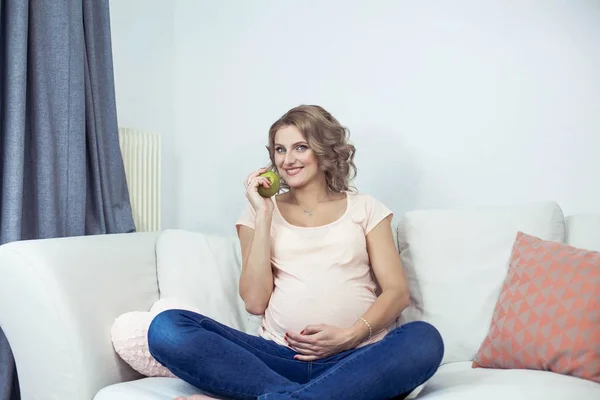Young beautiful pregnant woman is resting at home. Young pregnant woman is eating an apple. Health. Portrait of a beautiful pregnant woman. Cozy.