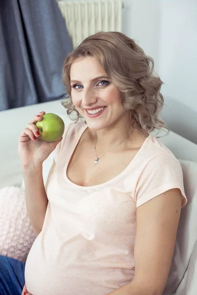 Giovane bella donna incinta sta riposando a casa. La giovane donna incinta sta mangiando una mela. Salute. Ritratto di una bella donna incinta. Accogliente . — Foto Stock