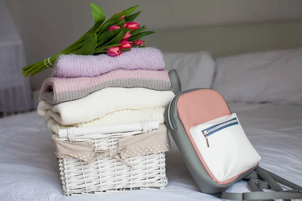 Un cesto con cose pulite sul letto. Accogliente. Maglioni lavorati a maglia, zaino, bouquet di tulipani. Vestiti primavera . — Foto Stock