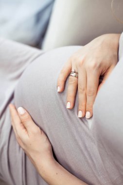 Genç, güzel ve hamile bir kadın kanepede oturuyor ve karnı dokunur. Çok güzel bir elbise içinde hamile. Annelik. Gebelik. Mutlu bir hamile kadın portresi. Rahat.