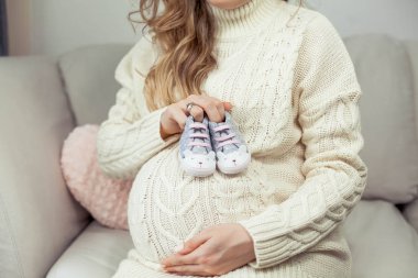 Genç, güzel ve hamile bir kadının karnı dokunur. Örme elbise hamile bir kadın kanepede oturuyor ve çocuk ayakkabıları tutar. Annelik. Gebelik. Mutlu bir hamile kadın portresi. Rahat.