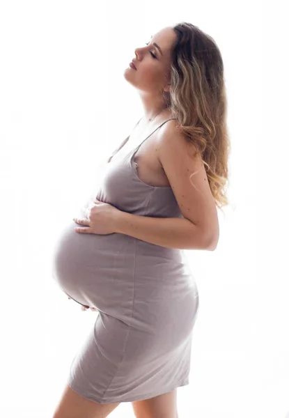 Uma jovem mulher grávida bonita está parada à janela. Grávida em um lindo vestido. Maternidade. Gravidez. Retrato de uma mulher grávida feliz. Aconchegante . — Fotografia de Stock