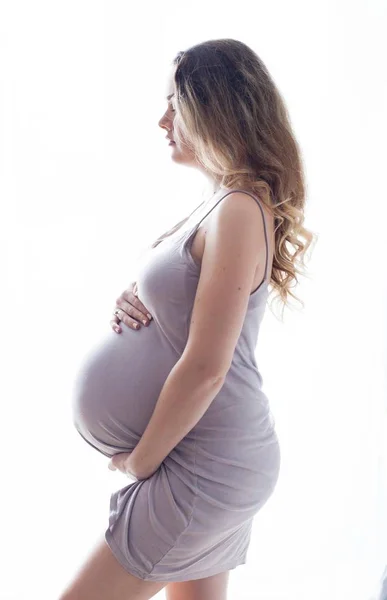 En ung vacker gravid kvinna står vid fönstret. Gravida i en vacker klänning. Moderskap. Graviditet. Porträtt av en glad gravid kvinna. Cozy. — Stockfoto