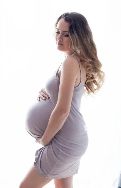 Una giovane bella donna incinta è in piedi vicino alla finestra. Incinta con un bel vestito. La maternità. Gravidanza. Ritratto di donna incinta felice. Accogliente . — Foto Stock