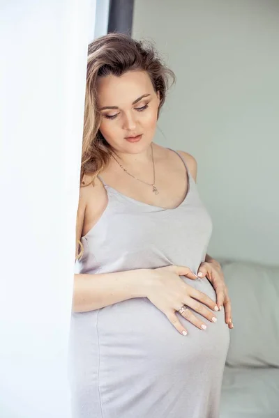 Μια νεαρή όμορφη έγκυος γυναίκα στέκεται δίπλα στο παράθυρο. Έγκυος σε ένα όμορφο φόρεμα. Η μητρότητα. Εγκυμοσύνη. Πορτρέτο της μια ευτυχισμένη γυναίκα έγκυος. Άνετο. — Φωτογραφία Αρχείου