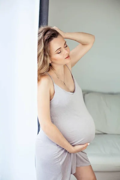 Μια νεαρή όμορφη έγκυος γυναίκα στέκεται δίπλα στο παράθυρο. Έγκυος σε ένα όμορφο φόρεμα. Η μητρότητα. Εγκυμοσύνη. Πορτρέτο της μια ευτυχισμένη γυναίκα έγκυος. Άνετο. — Φωτογραφία Αρχείου