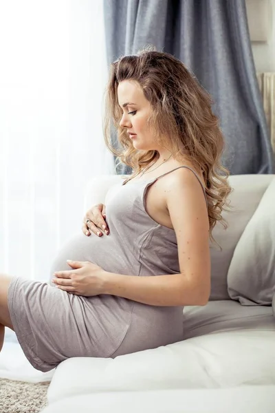 Una giovane donna incinta si siede su un divano e si tocca la pancia. Incinta con un bel vestito. La maternità. Gravidanza. Ritratto di donna incinta felice. Accogliente . — Foto Stock