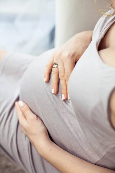 Młoda piękna kobieta w ciąży siedzi na kanapie i dotyka jej brzuch. W ciąży w piękną sukienkę. Macierzyństwo. Ciąży. Portret szczęśliwy kobiety w ciąży. Cozy. — Zdjęcie stockowe