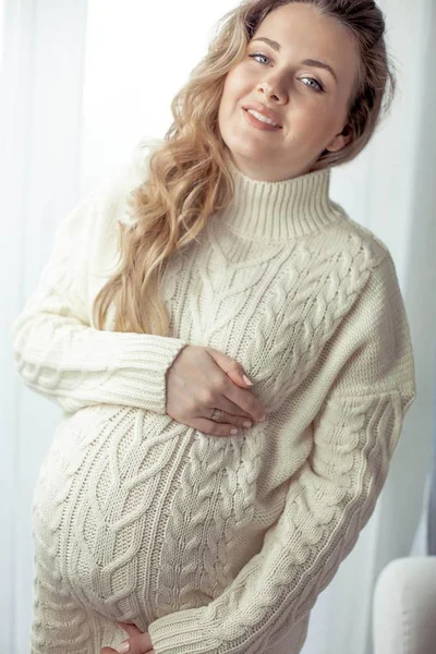 Een jonge mooie zwangere vrouw staat bij het raam. Zwanger in een gezellige gebreide jurk... Moederschap. Zwangerschap. Portret van een gelukkig zwangere vrouw. Gezellige. — Stockfoto