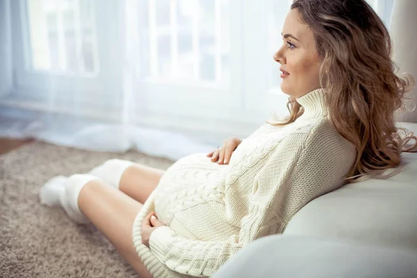 Uma jovem mulher grávida bonita está sentada à janela. Grávida em um vestido de malha acolhedor.. Maternidade. Gravidez. Retrato de uma mulher grávida feliz. Aconchegante . — Fotografia de Stock