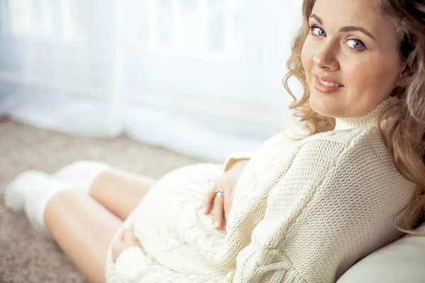 Uma jovem mulher grávida bonita está sentada à janela. Grávida em um vestido de malha acolhedor.. Maternidade. Gravidez. Retrato de uma mulher grávida feliz. Aconchegante . — Fotografia de Stock