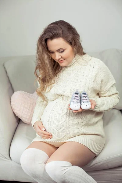 Μια νεαρή όμορφη έγκυος γυναίκα αγγίζει την κοιλιά της. Μια έγκυος γυναίκα σε ένα πλεκτό φόρεμα κάθεται στον καναπέ και κατέχει τα παπούτσια των παιδιών. Η μητρότητα. Εγκυμοσύνη. Πορτρέτο της μια ευτυχισμένη γυναίκα έγκυος. Άνετο. — Φωτογραφία Αρχείου