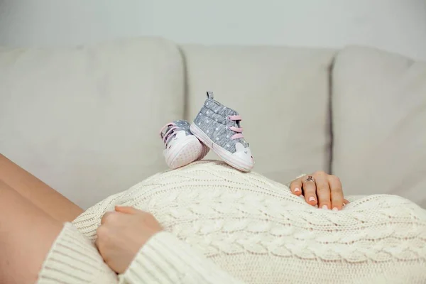 Una joven hermosa embarazada toca su barriga. Una mujer embarazada en un vestido de punto se encuentra en el sofá y sostiene los zapatos de los niños. Maternidad. Embarazo. Retrato de una mujer embarazada feliz. Acogedor . — Foto de Stock