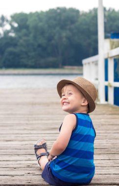 Sevimli küçük bir çocuk iskelede oturuyordu. Sahilde mutlu küçük çocuk. Yaz. Rekreasyon. Turizm. Deniz.