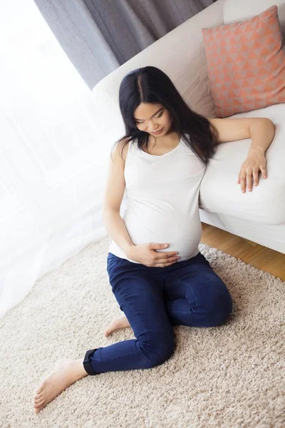 Piękna młoda Azjatka w ciąży kobieta siedzi przy oknie i dotyka jej brzuch. Ciąży. Zdrowia. Macierzyństwo. Przytulne portret kobiety w ciąży. Cozy. — Zdjęcie stockowe