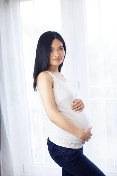 Bella giovane asiatica donna incinta in piedi vicino alla finestra e toccando la pancia. Gravidanza. Salute. La maternità. Un ritratto accogliente di una donna incinta. Accogliente . — Foto Stock
