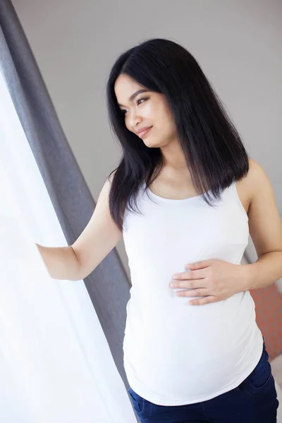 Bella giovane asiatica donna incinta in piedi vicino alla finestra e toccando la pancia. Gravidanza. Salute. La maternità. Un ritratto accogliente di una donna incinta. Accogliente . — Foto Stock