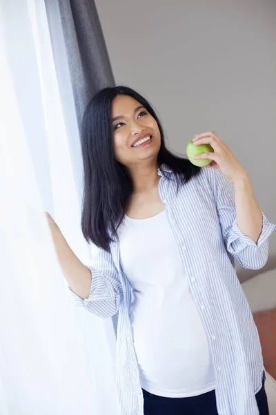 Mooie jonge Aziatische zwangere vrouw staat bij het raam en houdt van een appel. Zwangerschap. Gezondheid. Moederschap. Een gezellige portret van een zwangere vrouw. Gezellige. — Stockfoto