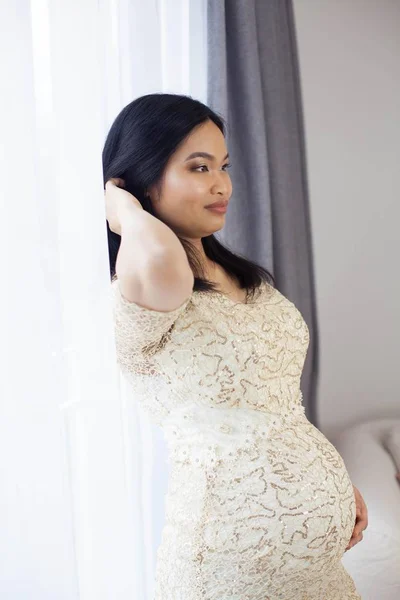 Linda jovem mulher grávida asiática em pé junto à janela e tocando sua barriga. Gravidez. Saúde. Maternidade. Um retrato acolhedor de uma mulher grávida. Aconchegante . — Fotografia de Stock