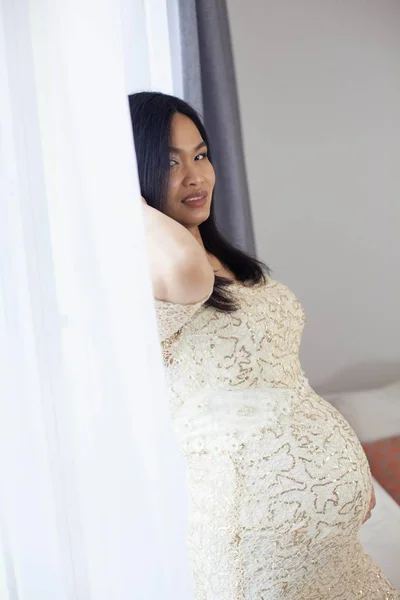 Linda jovem mulher grávida asiática em pé junto à janela e tocando sua barriga. Gravidez. Saúde. Maternidade. Um retrato acolhedor de uma mulher grávida. Aconchegante . — Fotografia de Stock