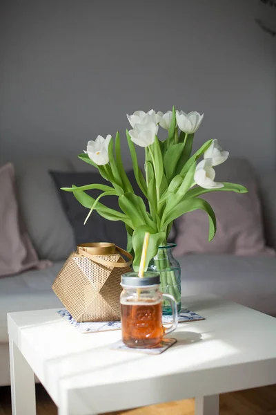 Interior. Room. A bouquet of tulips in a glass vase, a gold candlestick, a mug of lemonade on a white wooden table. Sofa, pillows. Spring. It's cozy. Living room. — Stock Photo, Image