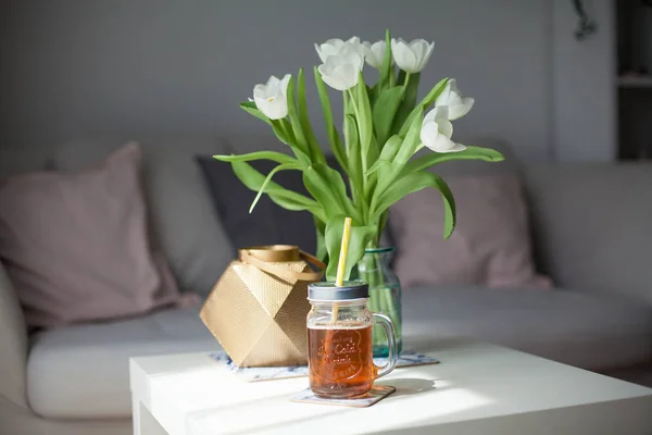 Interior. Quarto. Um buquê de tulipas em um vaso de vidro, um castiçal de ouro, uma caneca de limonada em uma mesa de madeira branca. Sofá, almofadas. Primavera. É aconchegante. Sala de estar . — Fotografia de Stock