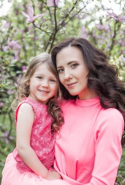 Küçük kızı olan genç güzel bir anne bahar park boyunca yürüyor. Bir anne ve çiçek açan magnolia yakınındaki küçük bir kız portresi. Bahar. Mutlu bir aile. Isı.