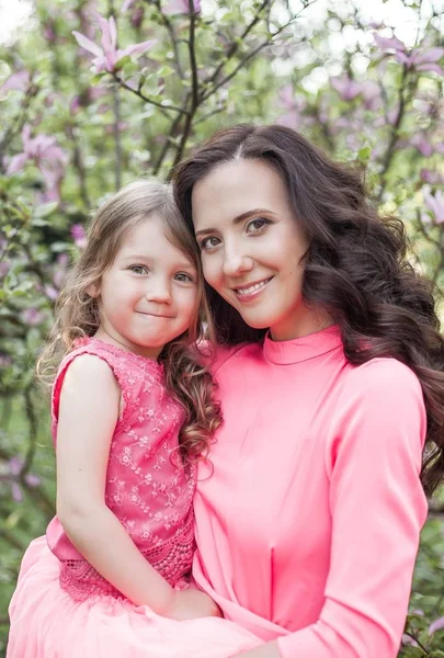 A young beautiful mother with a little daughter is walking along the spring park. Portrait of a mother and a little daughter near the blooming magnolia. Spring. A happy family. Heat. — Stock Photo, Image