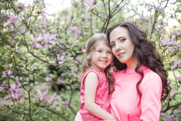 Молодая красивая мать с маленькой дочерью гуляет по весеннему парку. Портрет матери и маленькой дочери рядом с цветущей магнолией. Весной. Счастливая семья. Тепло . — стоковое фото