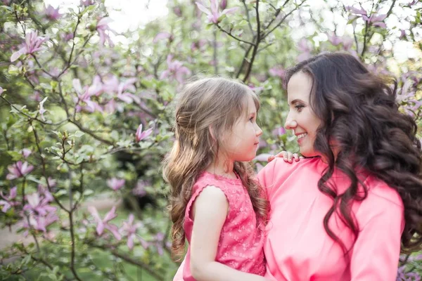 Seorang ibu muda yang cantik dengan seorang putri kecil berjalan di sepanjang taman musim semi. Potret seorang ibu dan seorang putri kecil di dekat magnolia yang sedang berkembang. Musim semi. Keluarga yang bahagia. Panas. . — Stok Foto