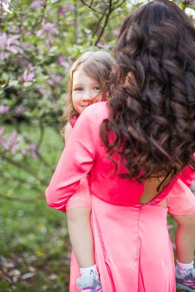 A young beautiful mother with a little daughter is walking along the spring park. Portrait of a mother and a little daughter near the blooming magnolia. Spring. A happy family. Heat. — Stock Photo, Image