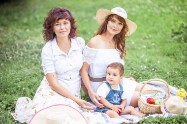 Genç bir anne, büyükanne ve küçük oğlu bir takas oynuyorlar. Piknik. Bahar. Yaz. Isı. Aile çimlerde oturuyor.