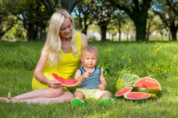 Piękna młoda matka synka siedząc na trawie i jedzenie arbuza. Piknik. Mama i syn są odpoczynku w parku. Rodzina. Szczęścia. Miłość. — Zdjęcie stockowe