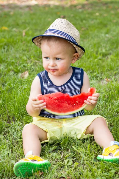A cute little boy is sitting on the grass and eating a watermelon. Picnic. The kid is resting in the park. Portrait of a little boy. A family. Happiness. Love. — Stock Photo, Image