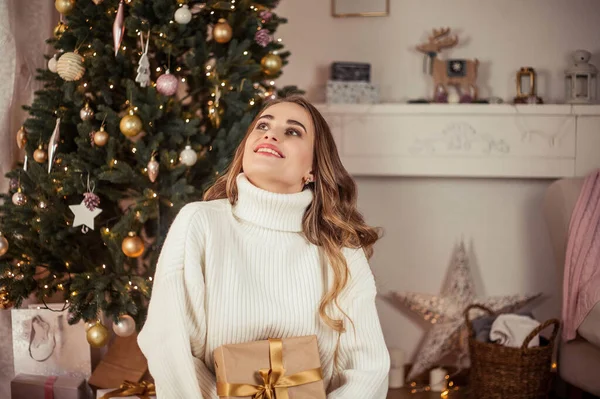 Uma bela jovem senta-se junto a uma árvore de Natal e abre presentes. No Natal. Ano Novo. Aconchegante . — Fotografia de Stock