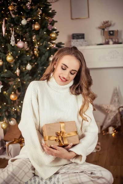 Eine schöne junge Frau sitzt an einem Weihnachtsbaum und öffnet Geschenke. Weihnachten. Neujahr. gemütlich. — Stockfoto