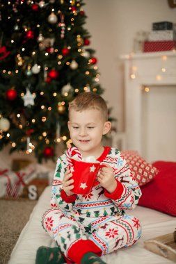 Noel pijamalı küçük bir çocuk Noel ağacının yanında oturuyor ve elinde bir kupa marşmelovlu kakao tutuyor. Noel zamanı. Yeni Yıl.