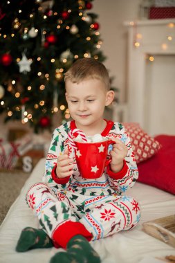 Noel pijamalı küçük bir çocuk Noel ağacının yanında oturuyor ve elinde bir kupa marşmelovlu kakao tutuyor. Noel zamanı. Yeni Yıl.