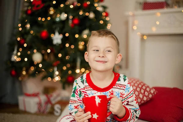 크리스마스 잠옷을 입은 한 소년 이 크리스마스 트리 근처에 앉아 마시멜로와 코코아 한 조각을 들고 있습니다. 크리스마스. 새해. — 스톡 사진
