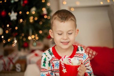 Noel pijamalı küçük bir çocuk Noel ağacının yanında oturuyor ve elinde bir kupa marşmelovlu kakao tutuyor. Noel zamanı. Yeni Yıl.