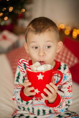 Noel pijamalı küçük bir çocuk Noel ağacının yanında oturuyor ve elinde bir kupa marşmelovlu kakao tutuyor. Noel zamanı. Yeni Yıl.