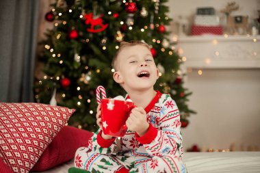 Noel pijamalı küçük bir çocuk Noel ağacının yanında oturuyor ve elinde bir kupa marşmelovlu kakao tutuyor. Noel zamanı. Yeni Yıl.