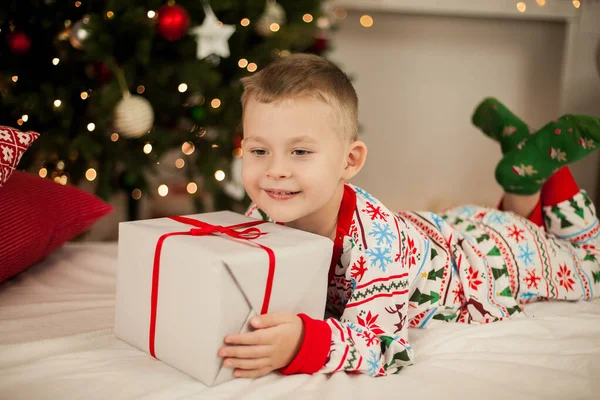 Malý chlapec v vánočním pyžamu sedí u vánočního stromku a otevírá dárky. Vánoce. Nový rok. — Stock fotografie