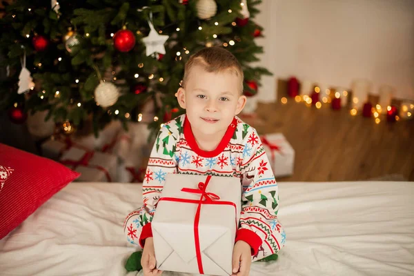 A little boy in Christmas pajamas sits near a Christmas tree and opens gifts. Christmas. New Year. — Stock Photo, Image