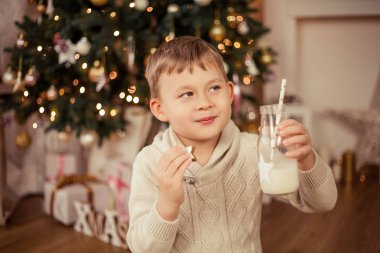 Küçük şirin çocuk bir Noel ağacının yanında oturur ve Noel kurabiyeleriyle süt içer. Noel zamanı. Yeni Yıl.